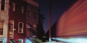  A freight train passes by row homes on the edge of Cherry Hill while the headlights of waiting cars shine through.Photography by Patrick Joust